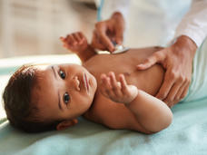 Photo of an infant lying on an exam table with an adult holding a stethoscope to the baby's chest.