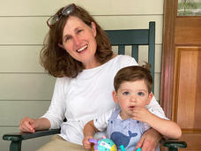 A smiling woman, Dr. Corinne Linardic, with red hair wearing a white shirt and a long, tan skirt sits in a chair with her arm around a child.
