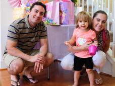 Joe and Erika smile and crouch next to their daughter, Melissa. Erika’s arm is around Melissa, who holds a small plastic bucket and smiles at the camera.