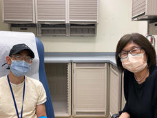 A doctor, Dr. Alice Chen, with short, dark hair wearing glasses and a surgical mask sits next to a patient wearing a hat, glasses, surgical mask, and yellow t-shirt in an exam room.