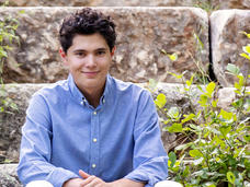 A young man, Callan, with brown hair and eyes, sits on a stone step wearing a blue button-up shirt and dress pants and smiles at the camera.