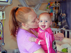 A woman, Kallie, with blond hair in a ponytail kisses the cheek of a smiling, blue-eyed baby, Sophie, wearing a pink superhero cape and facing the camera.