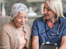Una paciente con un pañuelo en la cabeza se sienta junto a una médica quien le muestra a la paciente un dispositivo móvil.