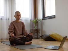 A middle-aged woman with a shaved head, sitting cross-legged on a yoga mat in front of a laptop computer.