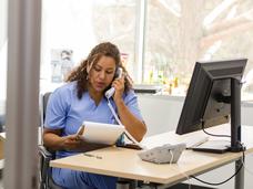 A woman in scrubs, sitting at a desk in a medical exam room, talks on a landline phone.