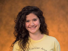 Juanita stands smiling at the camera, with dark brown, wavy hair and dark brown eyes, wearing a yellow T-shirt printed with “Behold Be Gold.”