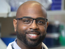 A photo of a man, Dr. Troy McEachron, who standing in a lab, smiling at the camera, and wearing glasses and a lab coat.
