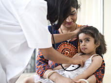 A health provider holds a stethoscope to a child's chest. The child sits on her mother's lap.