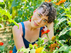 Hailey, with short, light-brown hair, stands in a field of sunflowers wearing a sleeveless blue top and yellow glasses and smiling at the camera.