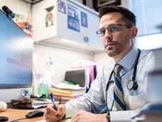 Dr. Matt Ehrhardt, with brown eyes and glasses, sits at a desk, pen in hand, wearing a dress shirt and tie and looking pensively into the distance.