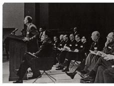 Black and white photo of U.S. Surgeon General Luther Terry addresses press at the release of the 1964 Report on Smoking and Health. Terry stands at a podium as staff and committee sit behind him. 