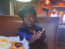 A boy, Cameron, wearing a ball cap and a black and blue striped T-shirt, sits in a restaurant booth and looks toward the camera.