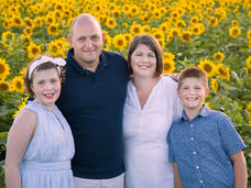 Girl in a blue dress with short hair held back with a headband, standing in a sunflower field with her family.