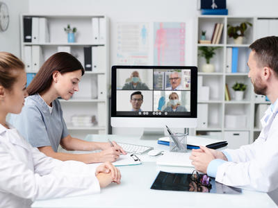 Three health care providers sit around a conference table having a videoconference with four health care providers working remotely. 