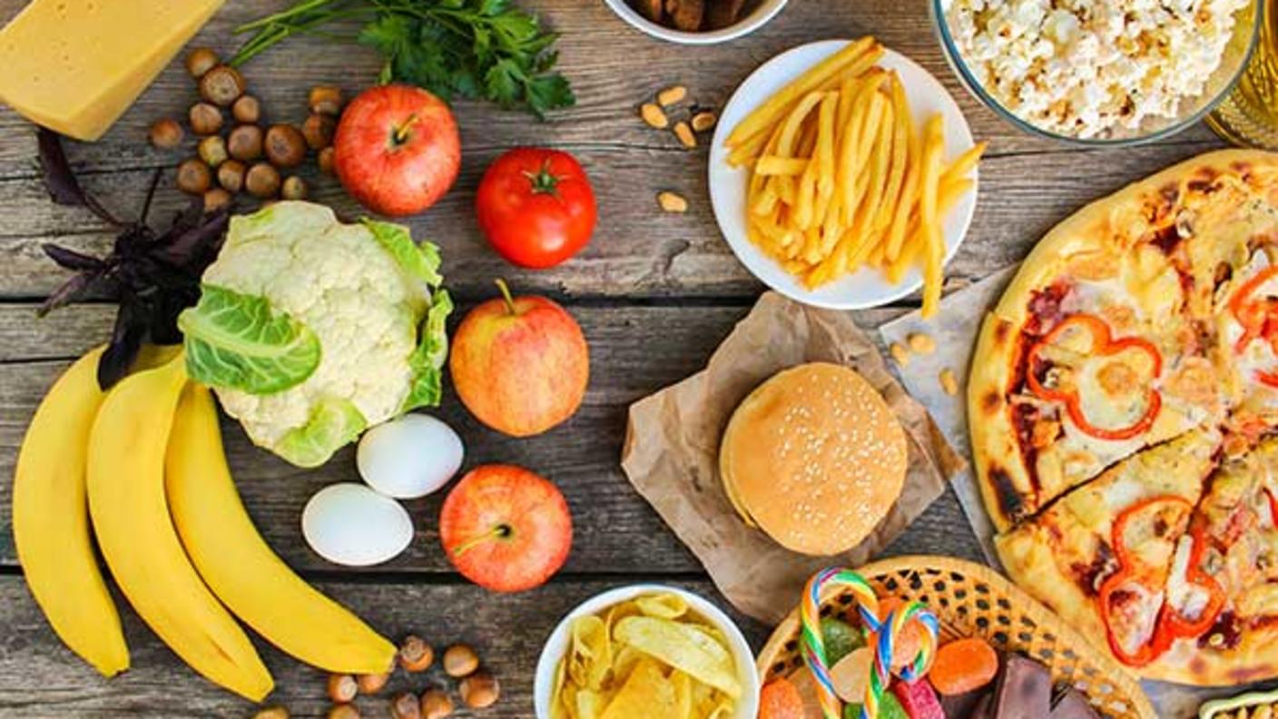 Overhead view of various foods arranged on wooden surface, including fresh produce like bananas, apples, and cauliflower on the left side, and processed foods like pizza, burgers, french fries, and snacks on the right side.