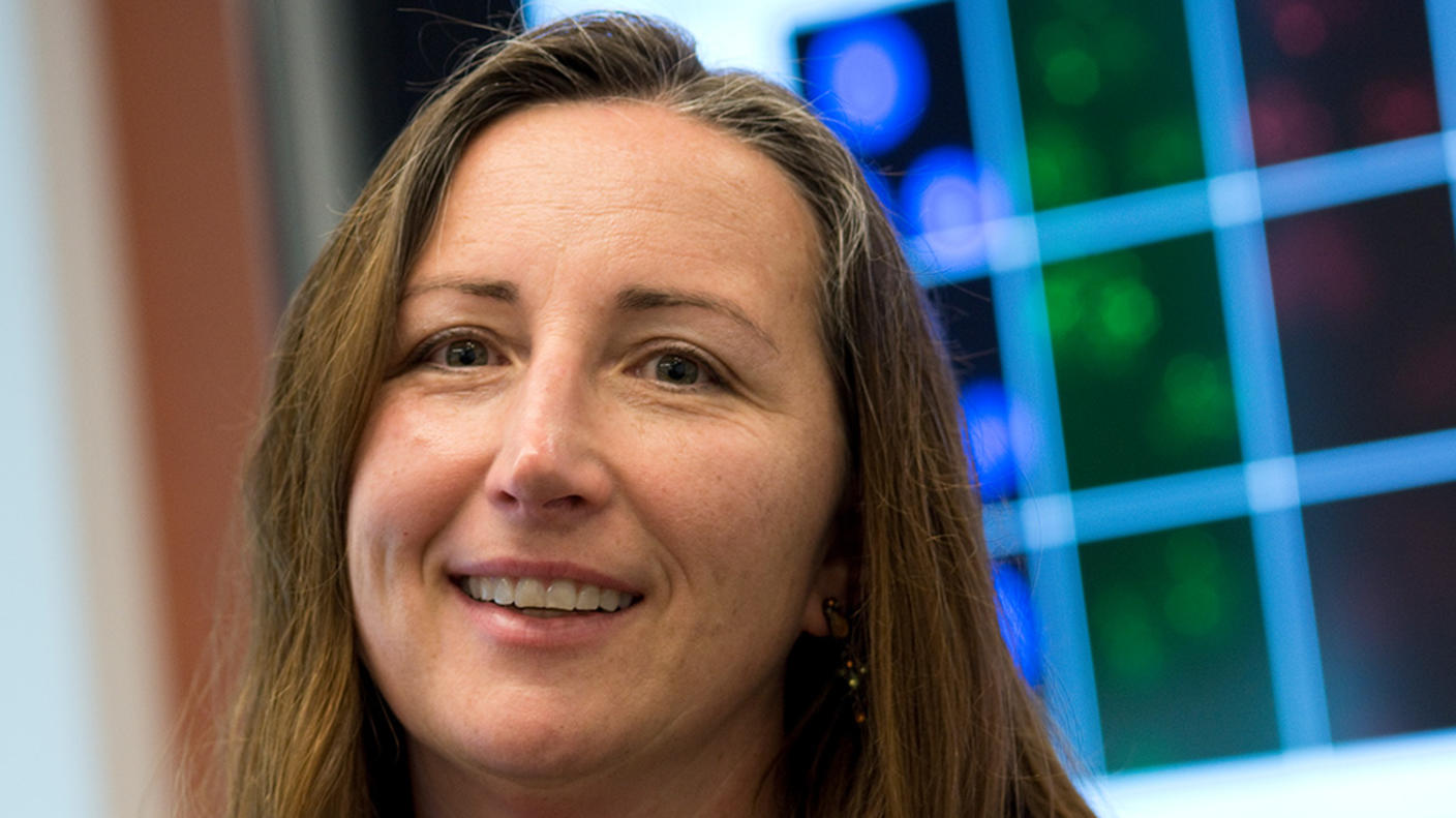 A White woman with light brown hair wearing a green blouse sitting in front of a large computer screen on the wall.