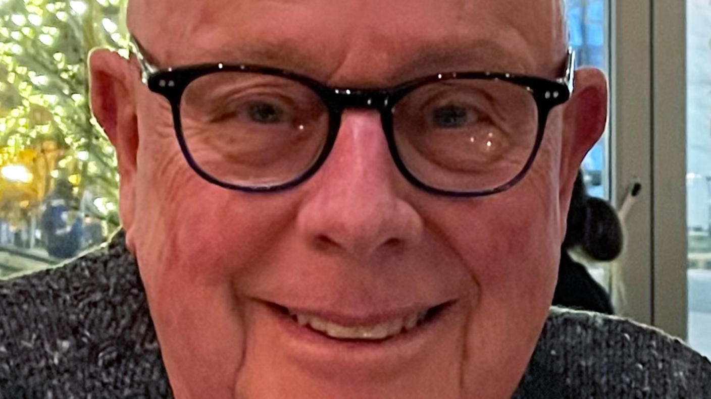 Man with glasses and grey knit sweater smiling at camera while sitting in front of glass doors. Outside are trees covered in white lights.