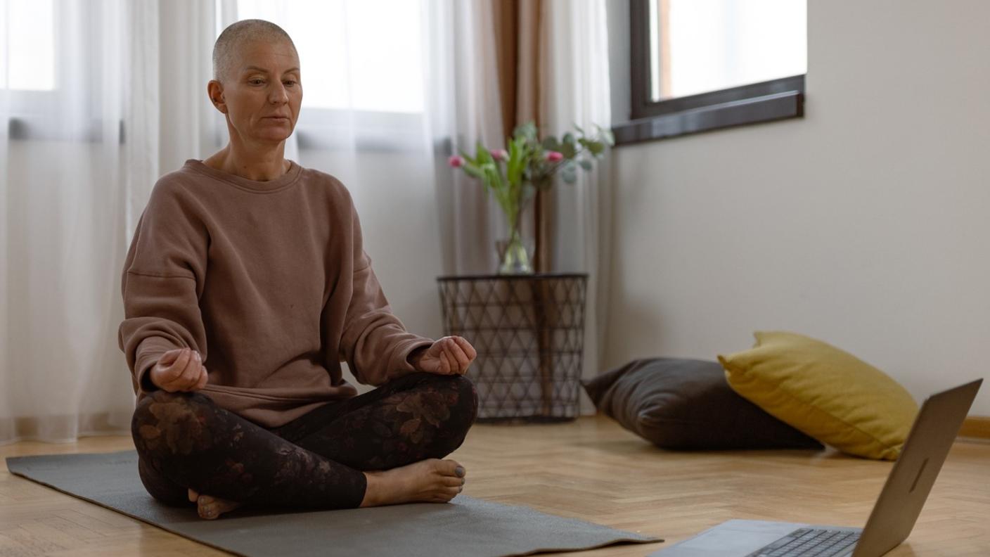 A middle-aged woman with a shaved head, sitting cross-legged on a yoga mat in front of a laptop computer.