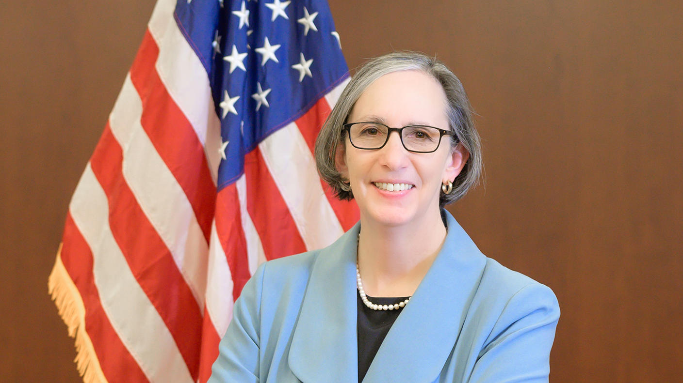La directora del Instituto Nacional del Cáncer, la doctora Rathmell, se para frente a la bandera de los EE. UU., sonriendo con los brazos cruzados.