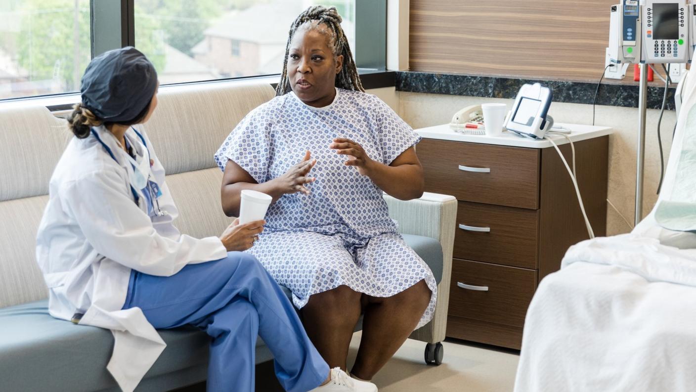 A middle-aged black woman in a hospital gown talking with a surgeon.