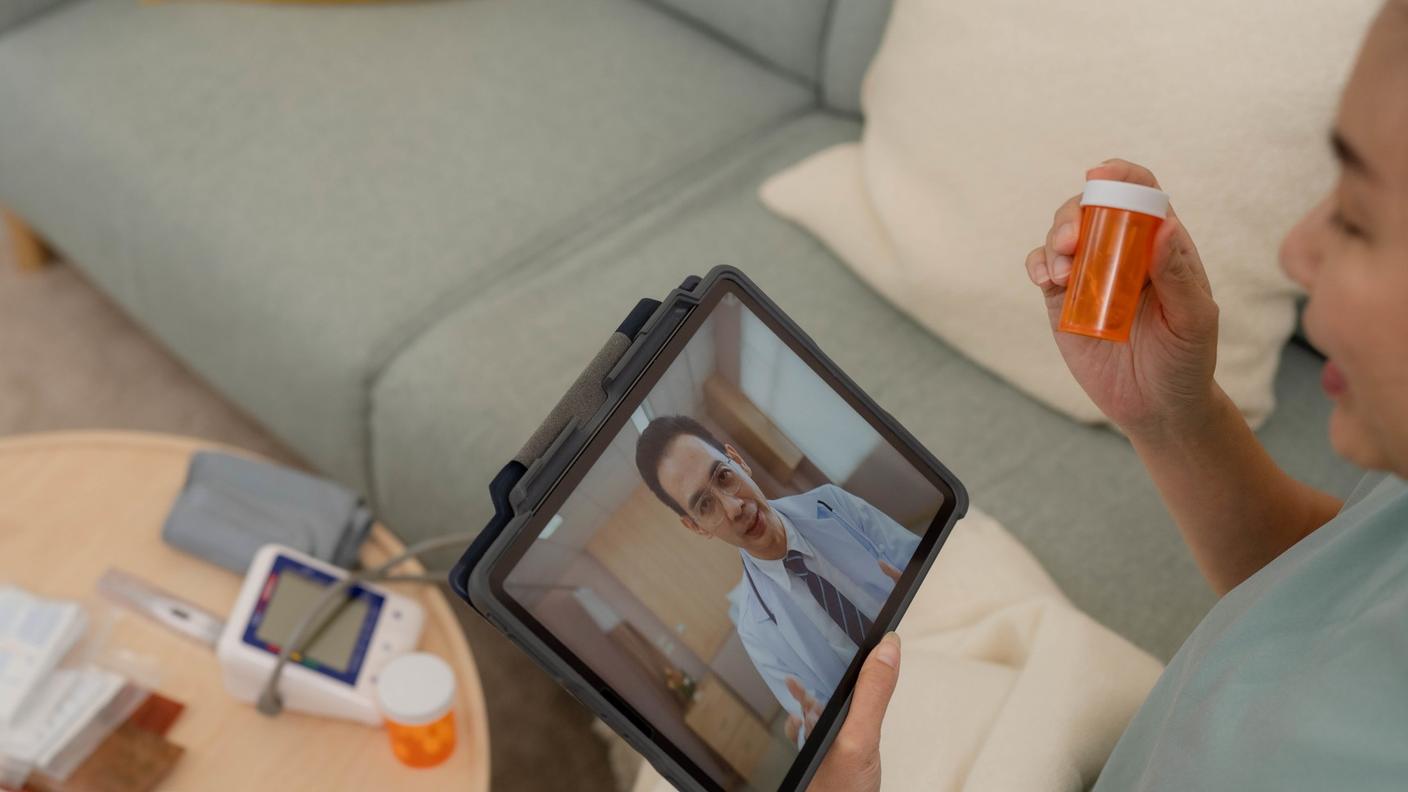 An Asian woman sitting on her couch, a pill bottle in her hand, talking to her doctor whose face can be seen on a tablet computer.