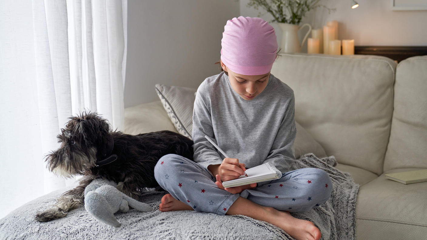 A child wearing a head scarf sits with legs crossed on a sofa, writing in a notebook as a dog sits beside the child.