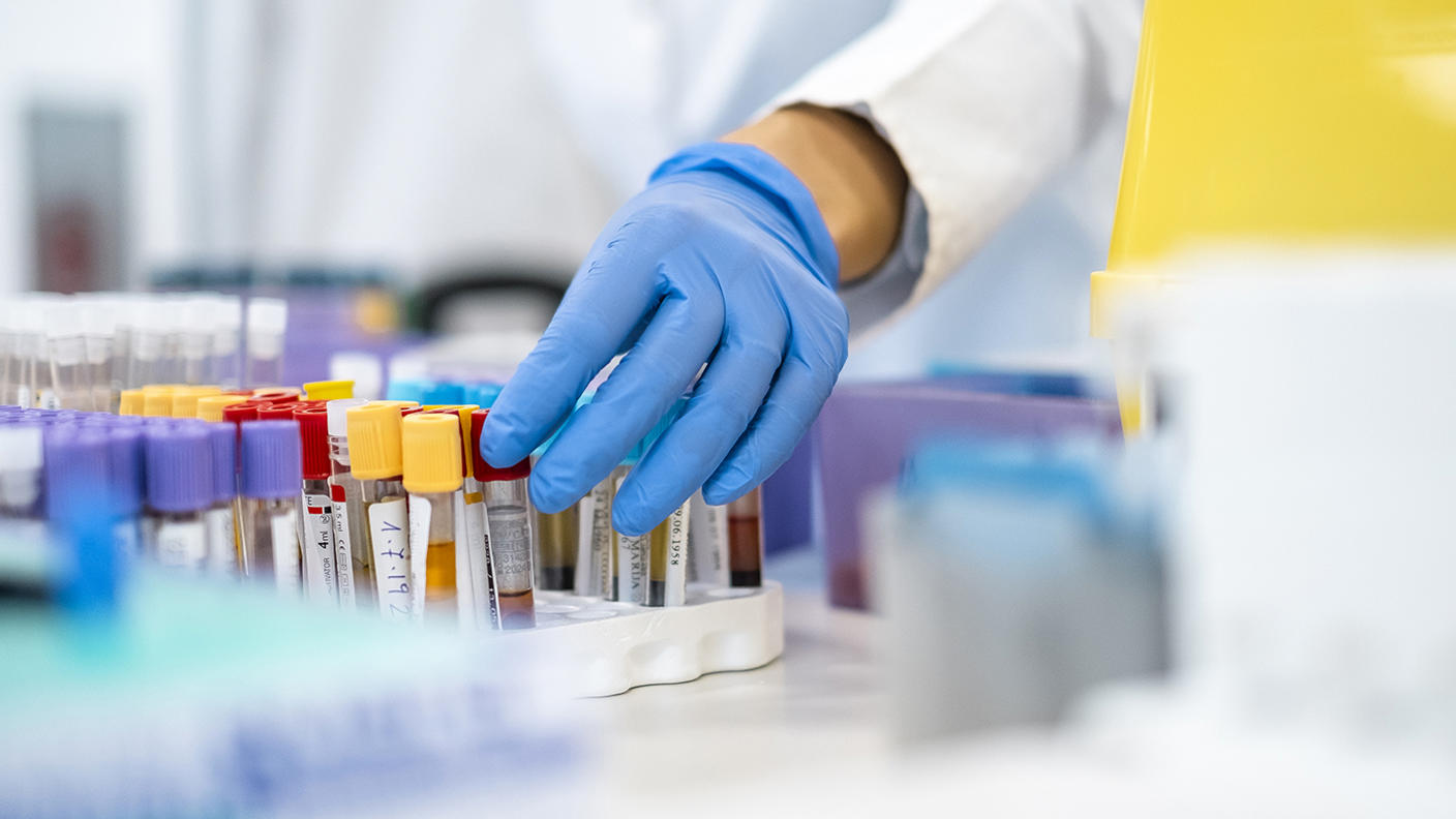 A gloved hand selects labeled blood sample tubes from a rack in a laboratory.
