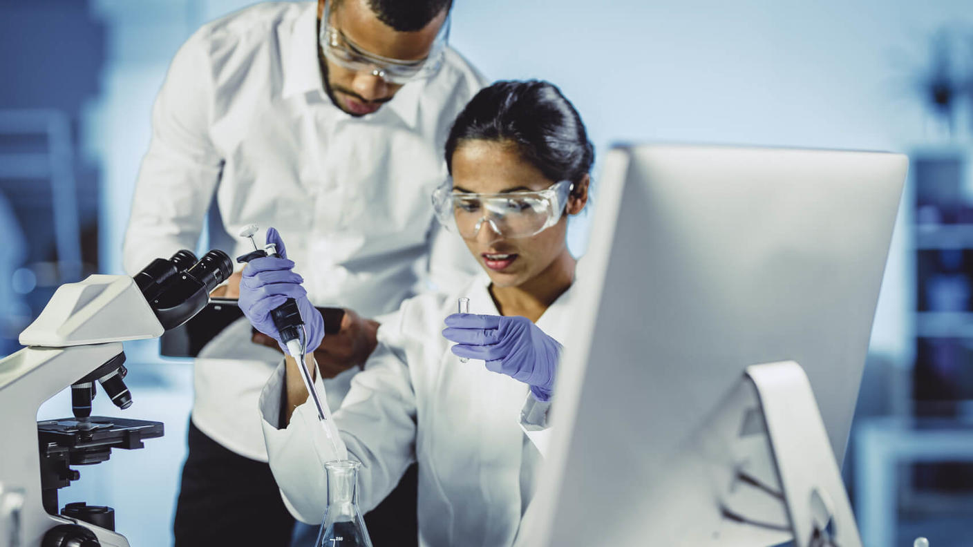 Researcher in white coat and goggles uses laboratory equipment while another researcher records observations.