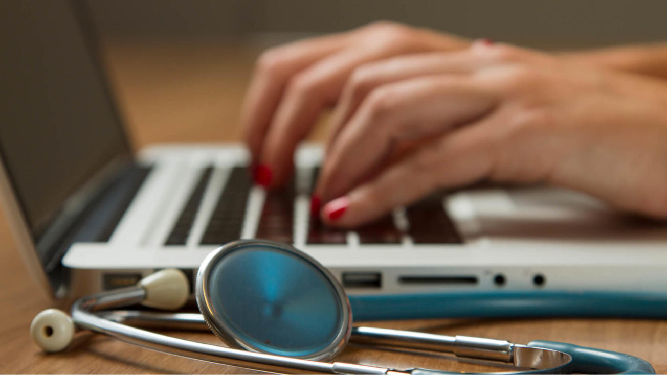 Medical professional typing on a computer with a stethoscope on the table.