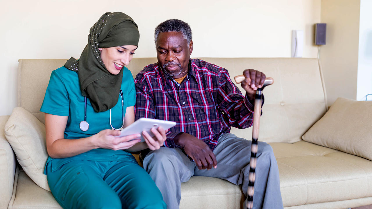 Healthcare worker in headscarf shows older Black male information on a tablet