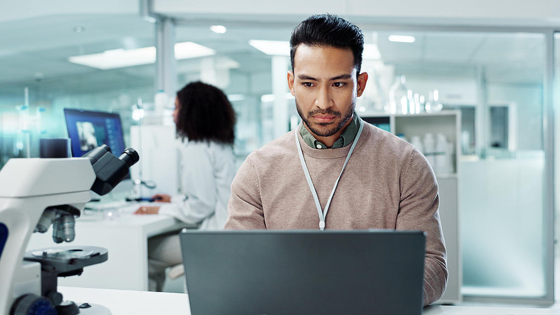 A man is working intently on a laptop in a modern laboratory. A microscope is visible in the foreground. In the background, a woman is seated at a desk, using a computer. The scene highlights ethnic and gender diversity in a professional scientific environment, with bright lighting and sleek lab equipment.