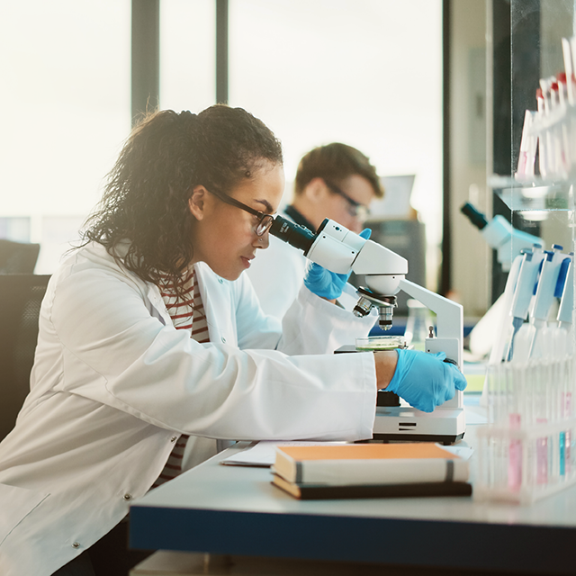 A young, female scientist in a lab coat and gloves examines a sample under a microscope in a laboratory, while two colleagues work in the background.
