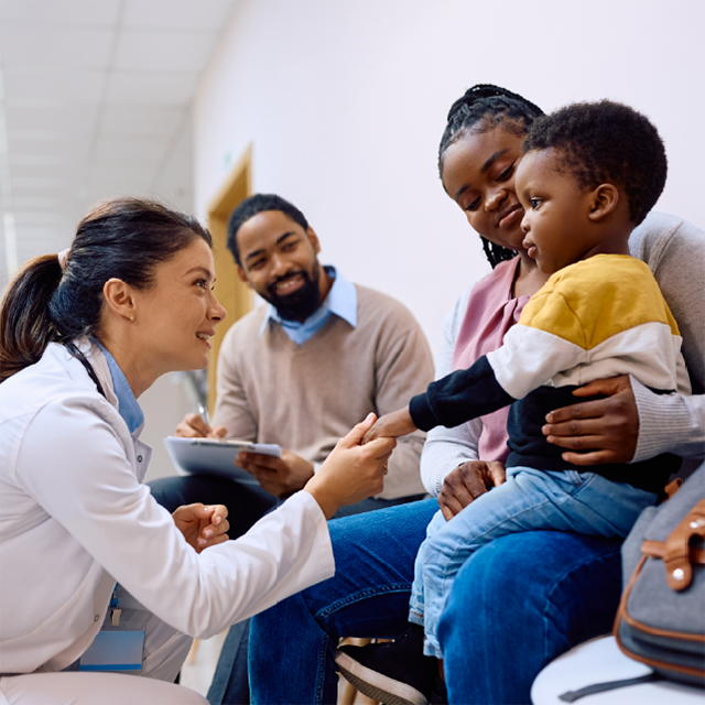 A health care provider smiles and holds the hand of a young child seated on a parent’s lap during a health care visit, while another adult smiles nearby.
