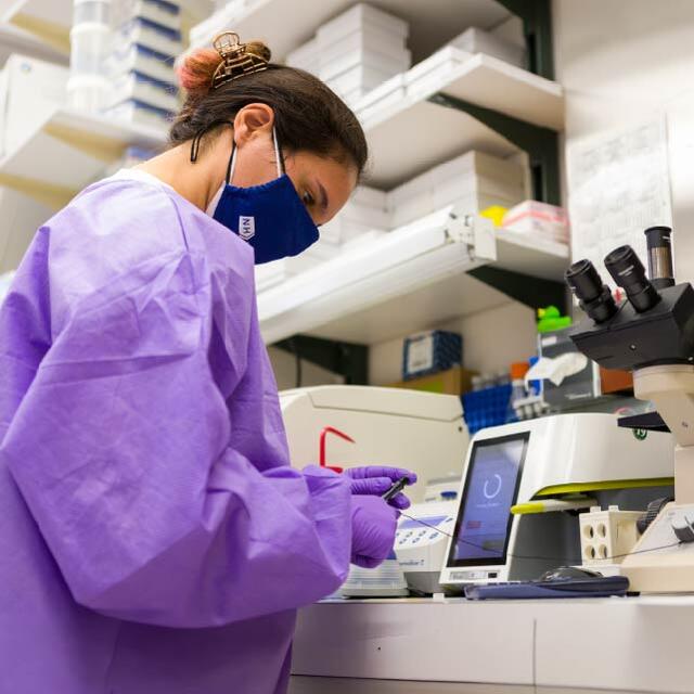 A researcher wearing protective clothing, gloves, and a face mask works beside a microscope and laboratory equipment.