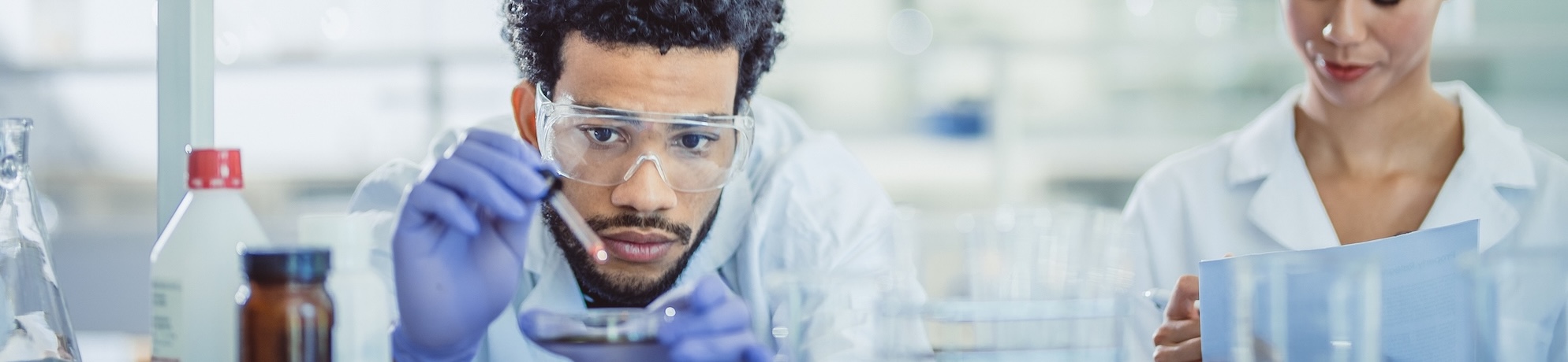 male scientist in a lab, wearing latex gloves and safety glasses, piping liquid into a small dish