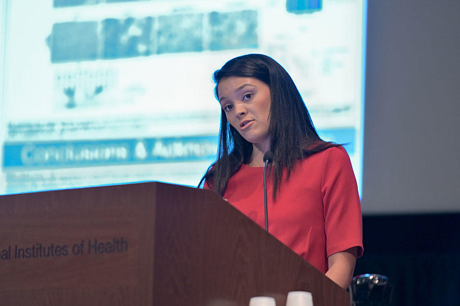 Sarah Martinez Roth stands speaking at a podium. She is wearing a red top and she has dark shoulder length hair. The podium is wood with the words “Institutes of Health” on it. In the background, out of a focus, a large projector screen with text and images on it.
