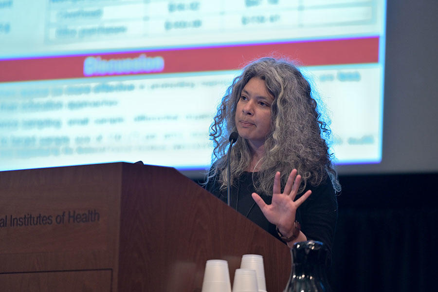 Yamilé Molina stands speaking at a podium. She is wearing a black top. The podium is wood with the words “Institutes of Health” on it. In the background, out of a focus, a large projector screen with text on it.
