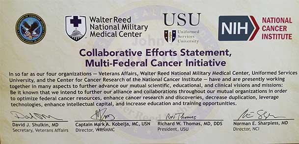 Enlarged copy of the memorandum titled "Collaborative Efforts Statement, Multi-Federal Cancer Initiative" displays logos from various organizations including the Department of Veterans Affairs, Walter Reed National Military Medical Center, Uniformed Services University, and National Cancer Institute. The text describes a commitment to collaboration in scientific, educational, and clinical efforts to enhance cancer research and resources. Signatures from officials of each organization are included at the bot