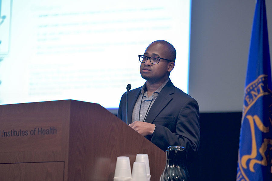 Dennis Jones stands speaking at a podium. He is wearing glasses and a suit jacket and button up shirt. The podium is wood with the words “Institutes of Health” on it. In the background, out of a focus, a large projector screen with text on it.