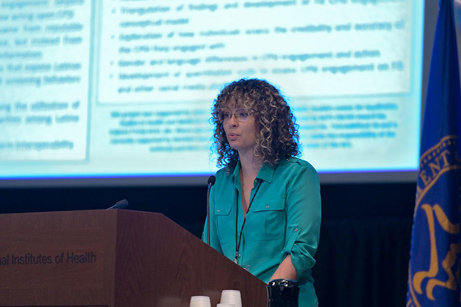Yonaira Rivera stands speaking at a podium. She is wearing glasses and a teal blouse. The podium is wood with the words “Institutes of Health” on it. In the background, out of a focus, a large projector screen with text on it.