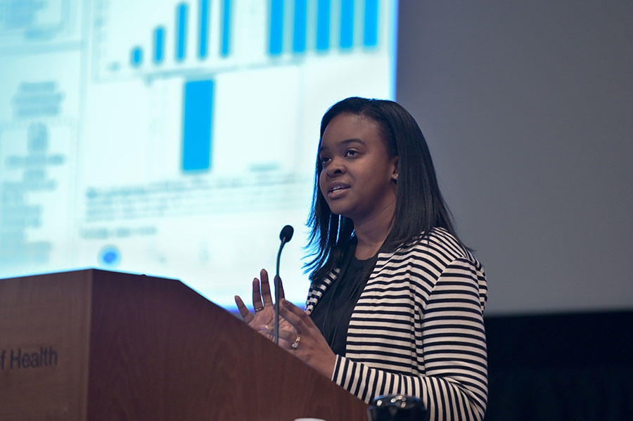 Lesley Chapman stands speaking at a podium. She is wearing a horizontally striped black and white top over a black shirt. The podium is wood with the word “ Health” on it. In the background, out of a focus, a large projector screen with text and charts on it.