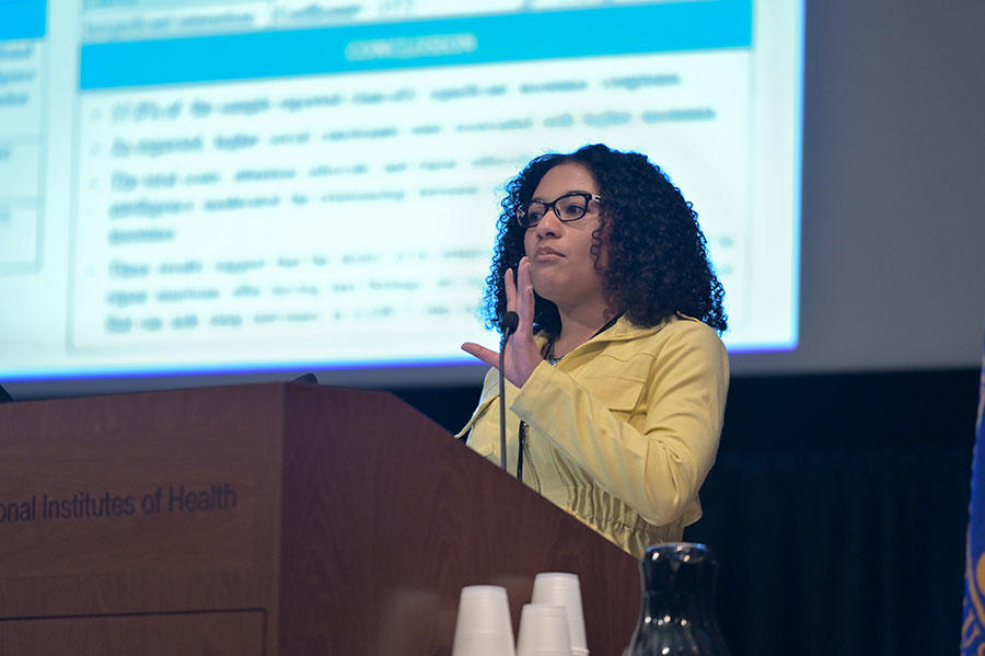 Chloe Martin stands speaking at a podium. She is wearing glasses and a yellow jacket. The podium is wood with the words “Institutes of Health” on it. In the background, out of a focus, a large projector screen with text on it.