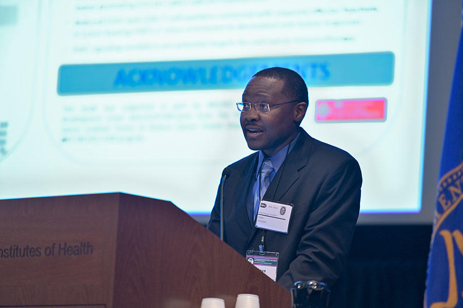 Steve Oghumu wears a suit and delivers an address at a podium. In the background, a blurry presentation slide can be seen. 