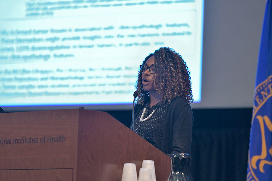 Adana Llanos stands speaking at a podium. She wearing glasses and a black long sleeve top over a polka dot blouse. The podium is wood with the words “Institutes of Health” on it. In the background, out of a focus, a large projector screen with text on it.