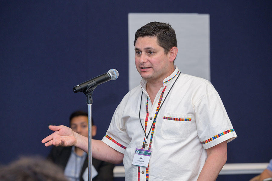 Daniel Liefwalker stands speaking into a microphone. He has short dark hair parted on the side and wears a white button up shirt with multi-colored fluting along the sleeves and center of the shirt.
