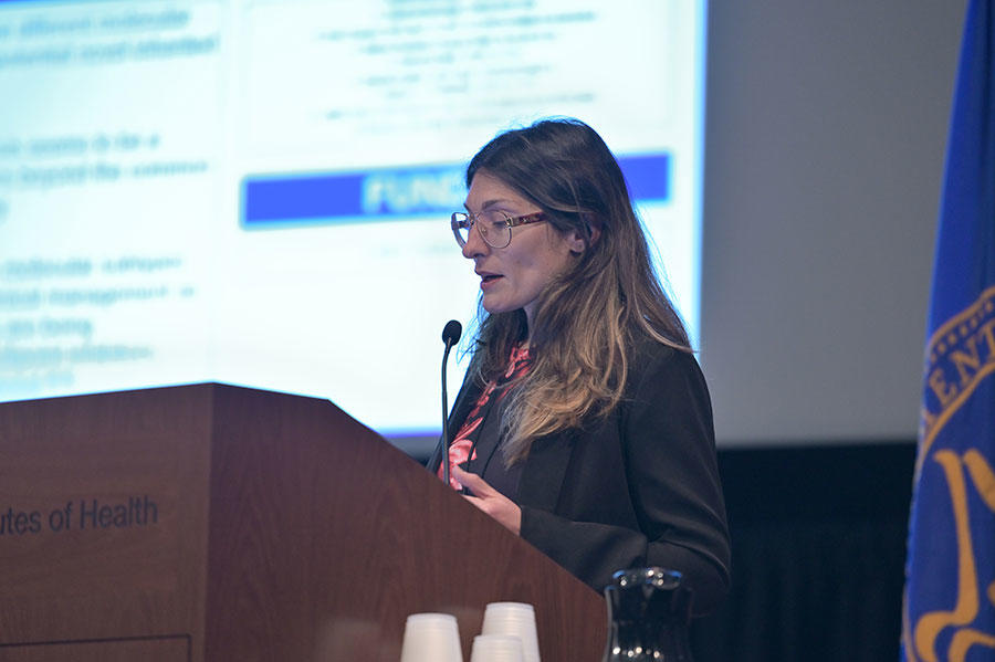 Rosa Munoz Xicola stands speaking at a podium. She wearing glasses and a black long sleeve top over a black-and-floral blouse. The podium is wood with the words “of Health” on it. In the background, out of a focus, a large projector screen with text on it.