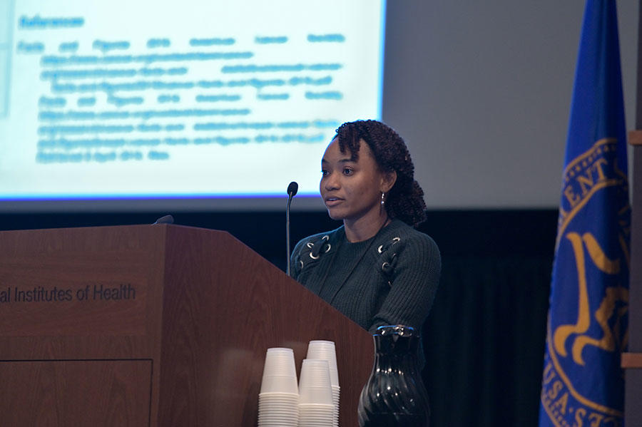 Carla Gibbs stands speaking at a podium. She is wearing a long sleeve black top. The podium is wood with the words “Institutes of Health” on it. In the background, out of a focus, a large projector screen with text on it.