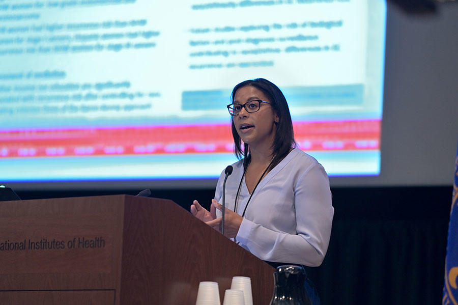 Ashley Felix stands speaking at a podium. She is wearing a long sleeve lilac colored top. The podium is wood with the words “Institutes of Health” on it. In the background, out of a focus, a large projector screen with text on it.