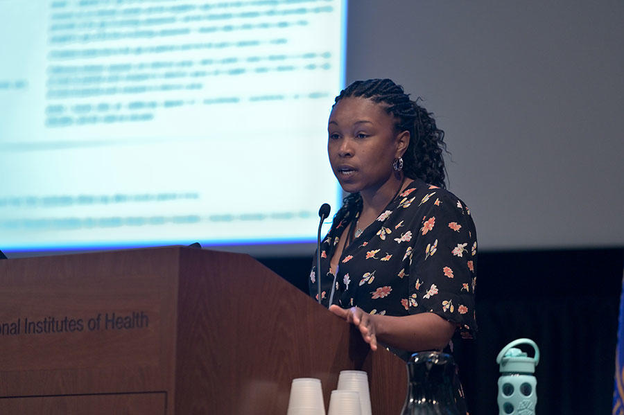 Jasmine McDonald stands speaking at a podium. She is wearing a short sleeve black top with differently sized flowers on it. The podium is wood with the words “Institutes of Health” on it. In the background, out of a focus, a large projector screen with text on it.