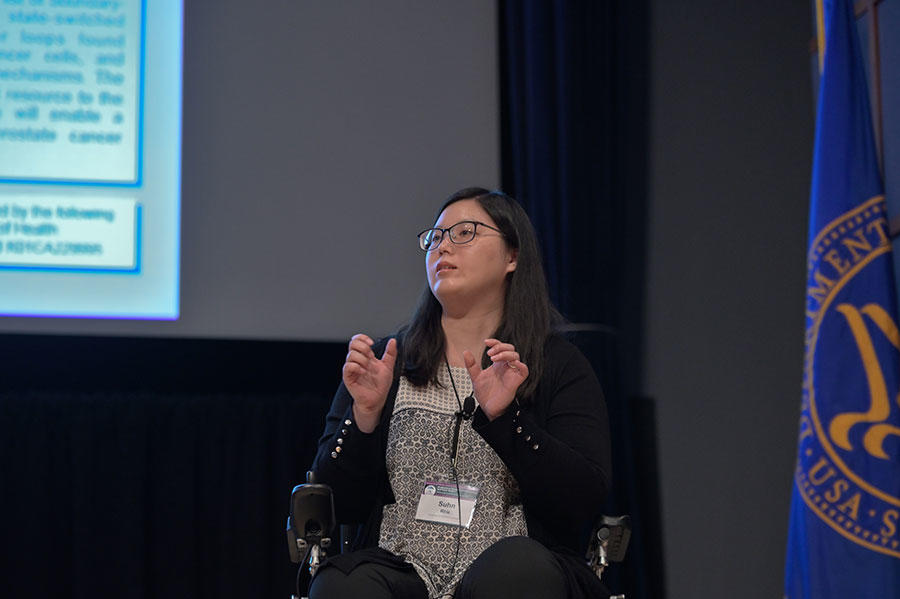 Suhn Rhie, in business dress wearing glasses and in a wheelchair, speaks on a stage. She is an Asian woman with shoulder length dark hair. On the background is a blue flag and part of a projector screen.